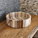 Switch A round Norhew vessel sink crafted from polished multi-colored banded onyx with distinct horizontal layers of cream, brown, and tan. 3 image