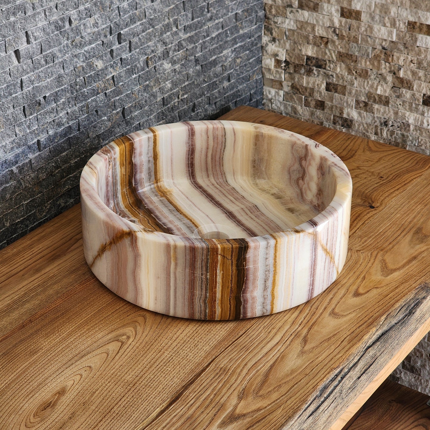A round Norhew vessel sink crafted from polished multi-colored banded onyx with distinct horizontal layers of cream, brown, and tan.
