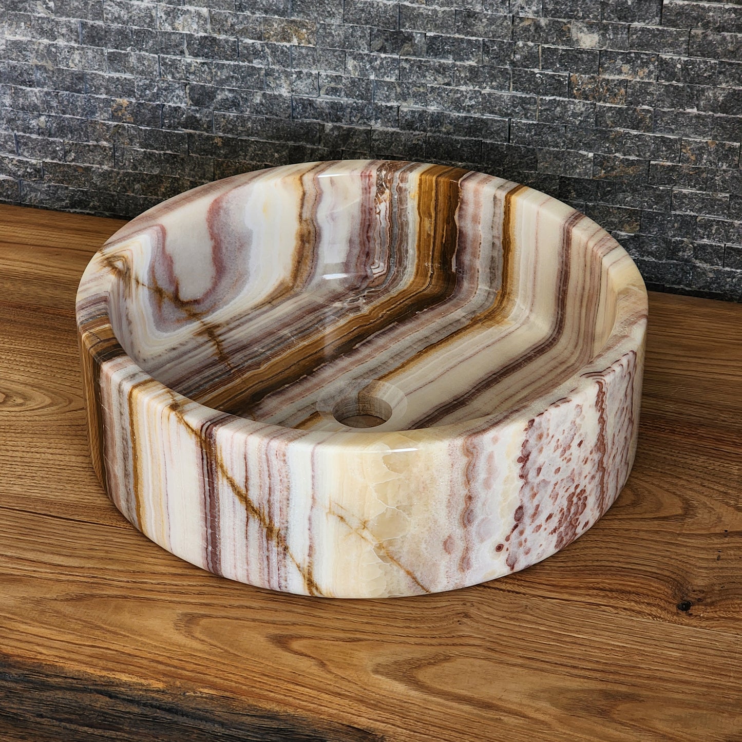 A round Norhew vessel sink crafted from polished multi-colored banded onyx with distinct horizontal layers of cream, brown, and tan.