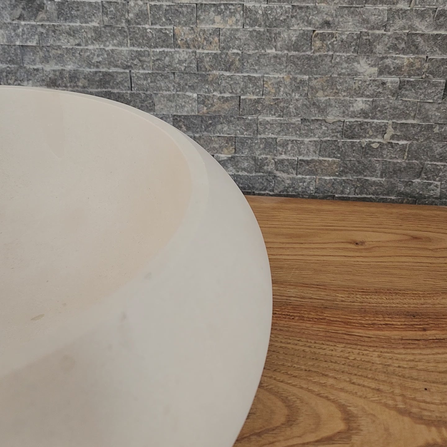A round Norhew cream limestone vessel sink with a soft matte finish and organic pebble-like shape on a natural wood vanity.