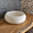 Switch A round Norhew cream limestone vessel sink with a soft matte finish and organic pebble-like shape on a natural wood vanity. 3 image