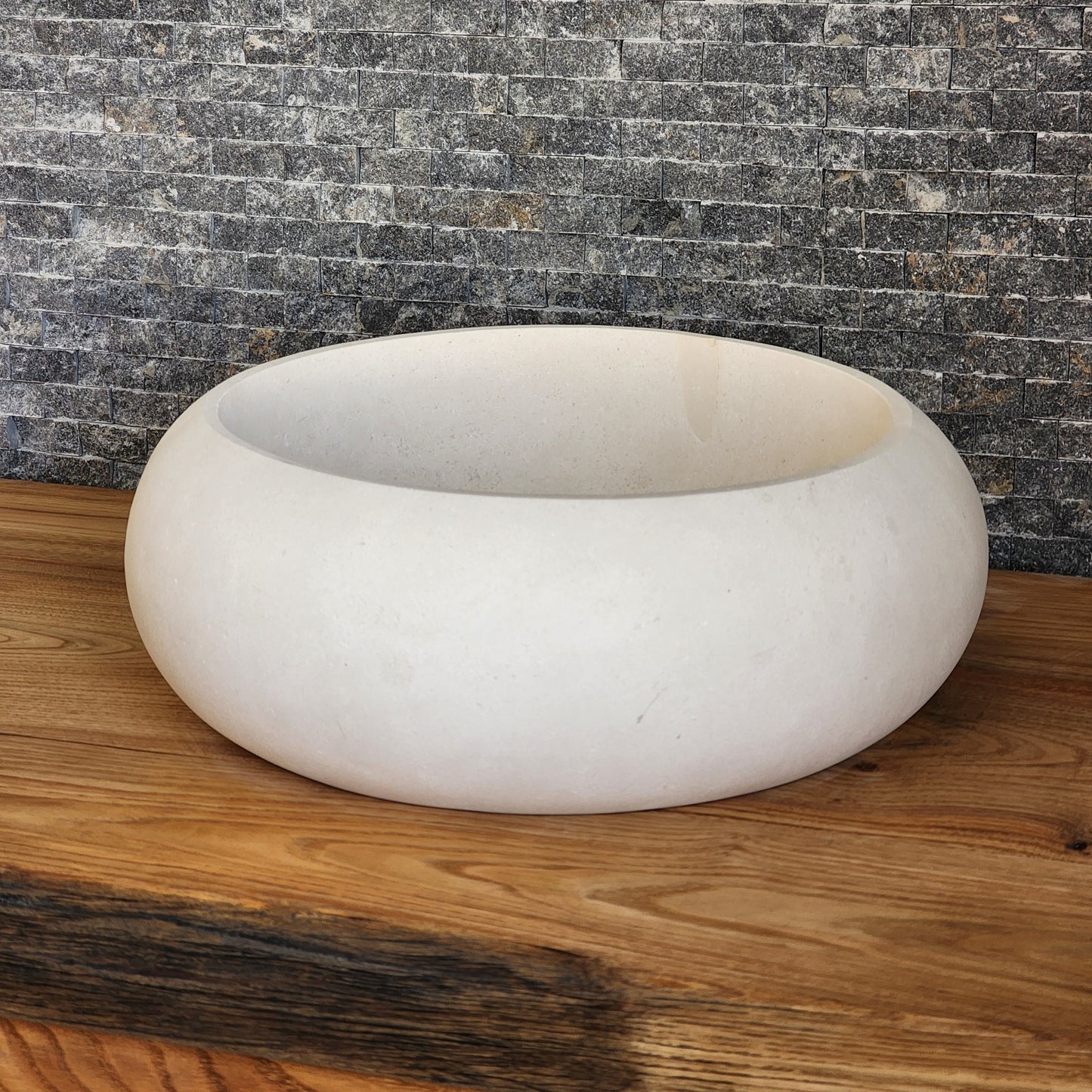 A round Norhew cream limestone vessel sink with a soft matte finish and organic pebble-like shape on a natural wood vanity.