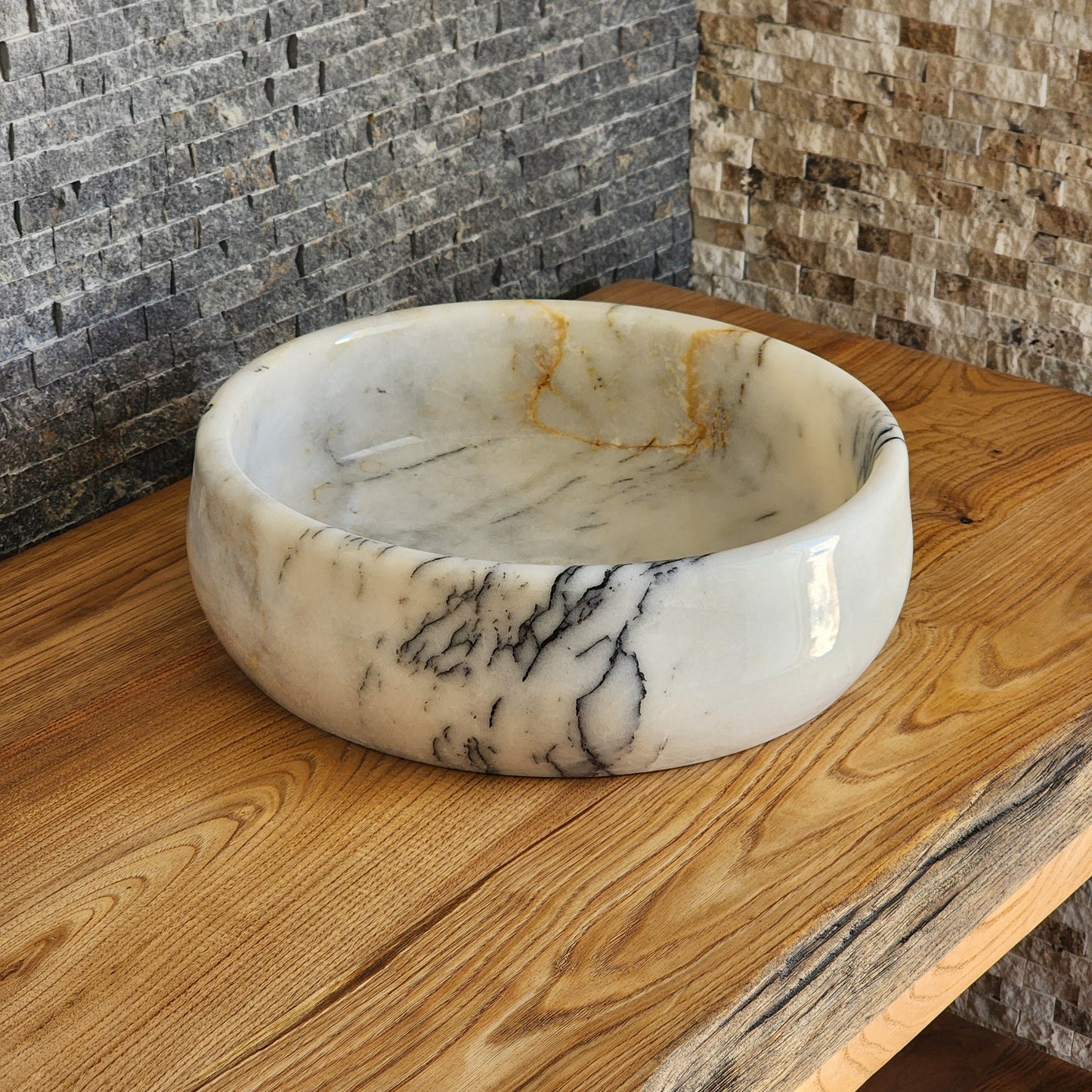 A round, hand-carved Norhew white marble vessel sink with distinct grey and gold veining, polished to a high-gloss finish on a solid wood countertop.