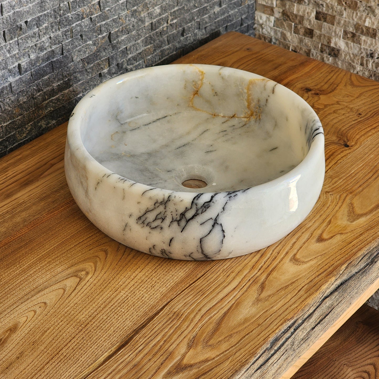 A round, hand-carved Norhew white marble vessel sink with distinct grey and gold veining, polished to a high-gloss finish on a solid wood countertop.