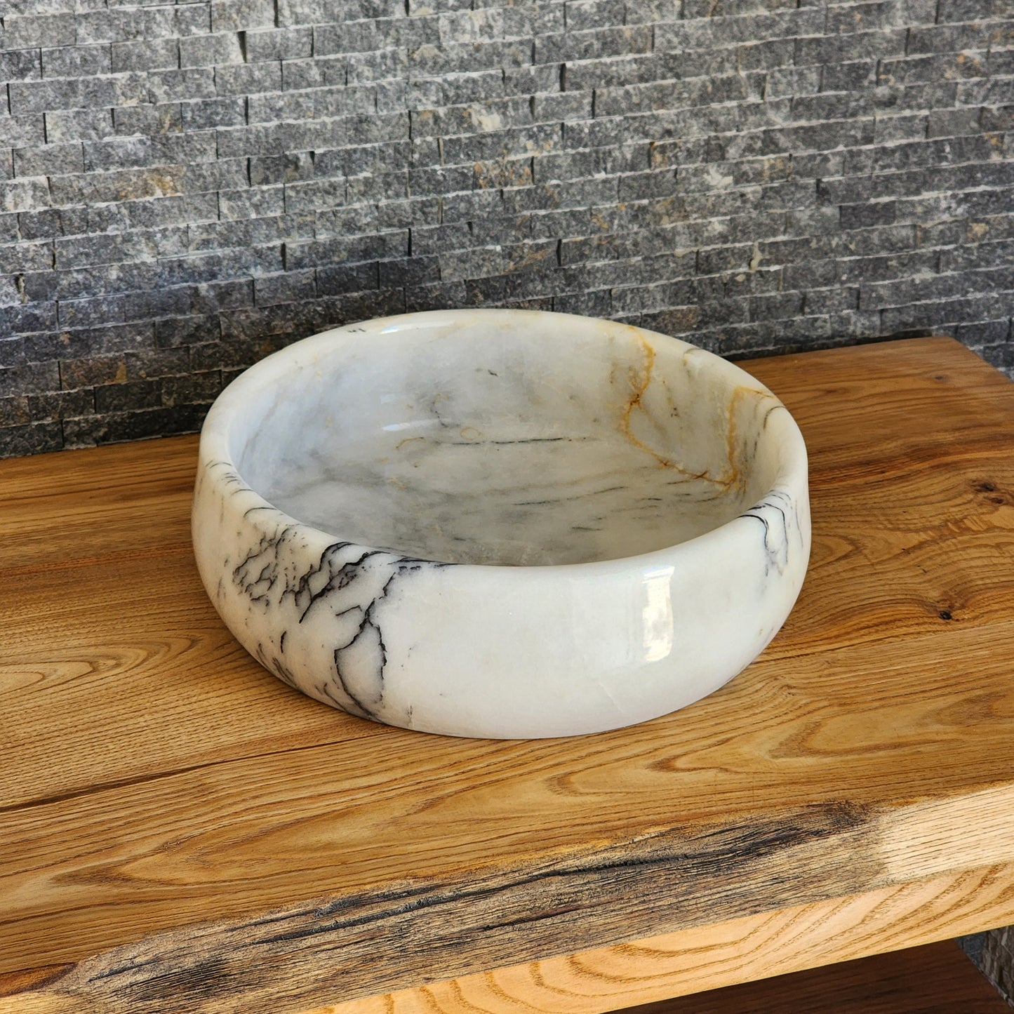 A round, hand-carved Norhew white marble vessel sink with distinct grey and gold veining, polished to a high-gloss finish on a solid wood countertop.