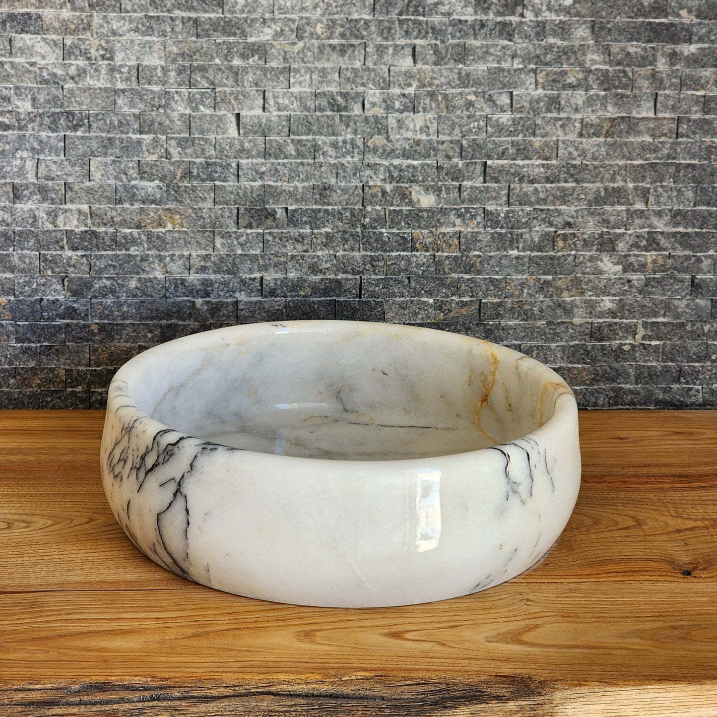 A round, hand-carved Norhew white marble vessel sink with distinct grey and gold veining, polished to a high-gloss finish on a solid wood countertop.
