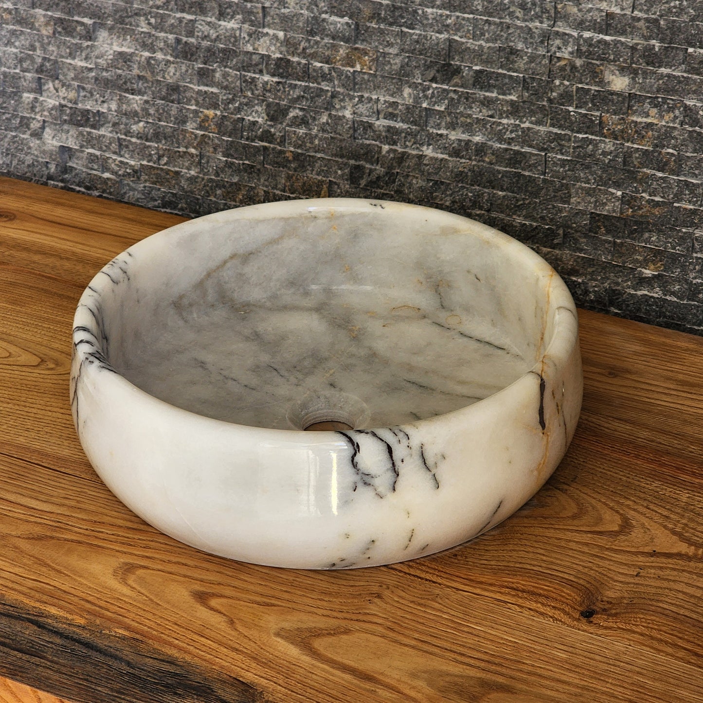 A round, hand-carved Norhew white marble vessel sink with distinct grey and gold veining, polished to a high-gloss finish on a solid wood countertop.